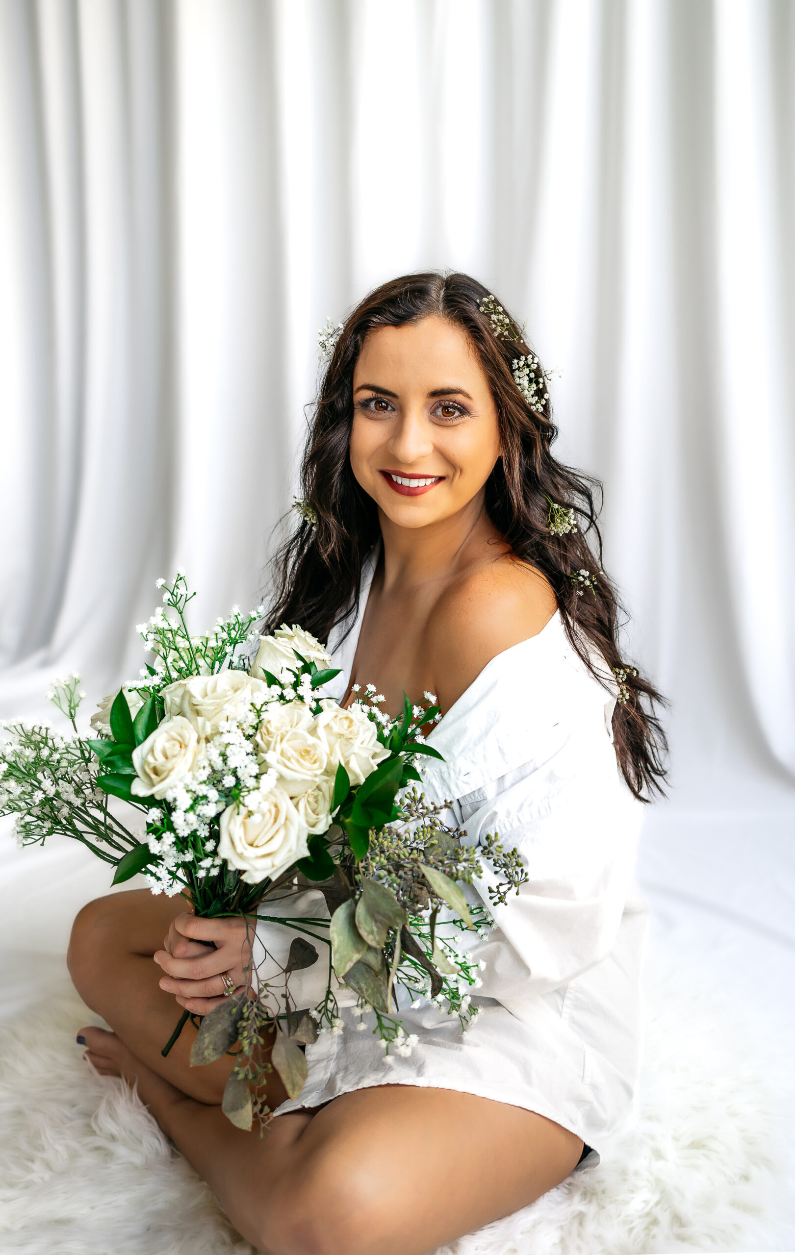 a 30th birthday photoshoot. client is wearing a white oversized loose fitting button down shirt with with her hair in soft curls with baby's breath flowers delicately placed throughout. white backdrop at-home session